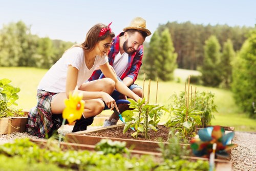 A man and a young girl are working together in a well-maintained garden, surrounded by lush greenery and trees in the background. They are tending to vegetable garden beds made of wooden borders, with visible plants growing in neat, organized rows. The garden beds contain leafy greens, herbs, and possibly young vegetable plants, with rich, dark soil. The foreground features a gravel pathway alongside the garden beds, leading through the outdoor space. The scene appears to be set on a bright, clear day with natural sunlight illuminating the plants and the two gardeners. This outdoor setting is indicative of a suburban garden in the Shortlands area, demonstrating typical gardening activities such as planting or inspecting crops, and reflects the kind of outdoor maintenance and gardening services provided by companies like Hedge Trimming Shortlands, enhancing the natural beauty and productivity of residential outdoor spaces.