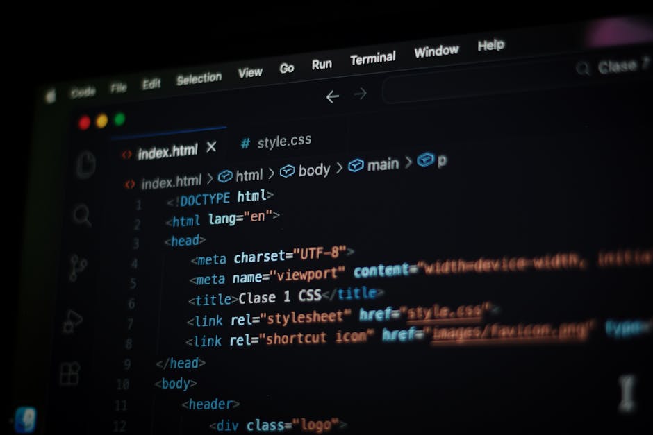 A person working indoors at a desk with dual computer monitors displaying lines of code and programming interfaces. The individual has short brown hair, a beard, and wears glasses and a gray sweater. 